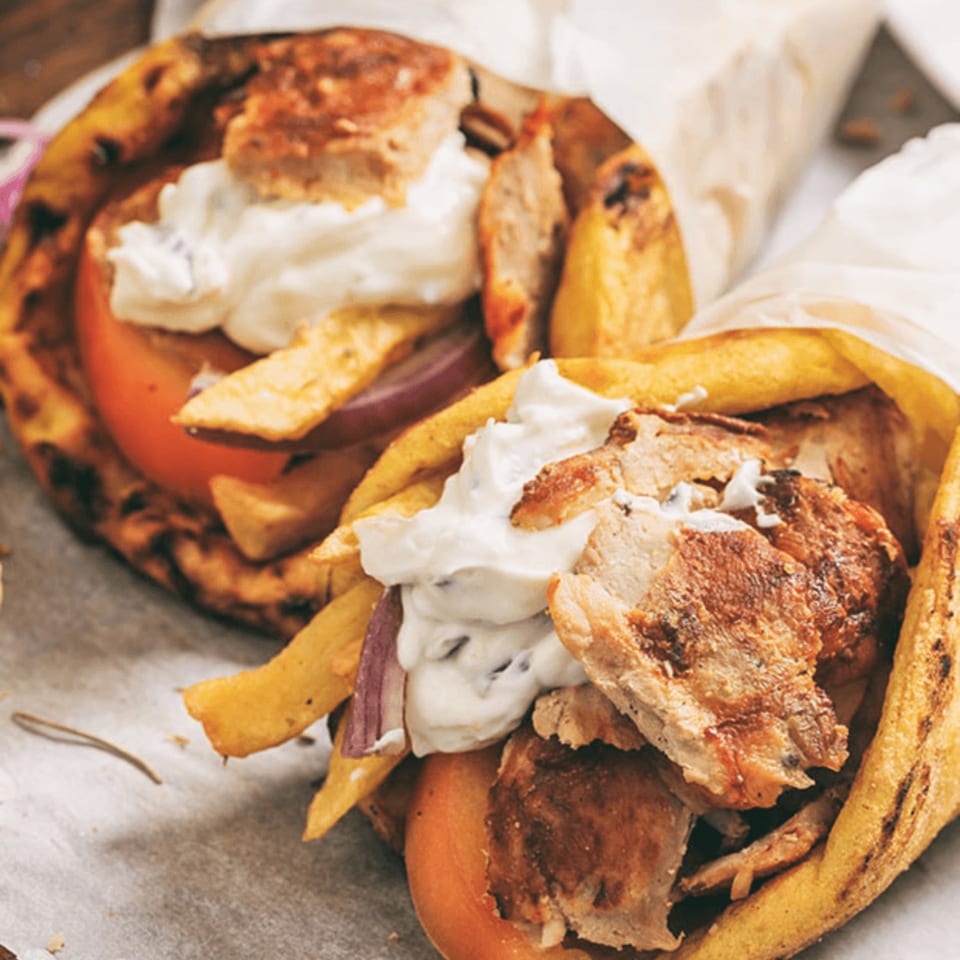 Two Greek gyros with chicken, fries, tomatoes, onions, and tzatziki sauce in pita bread.