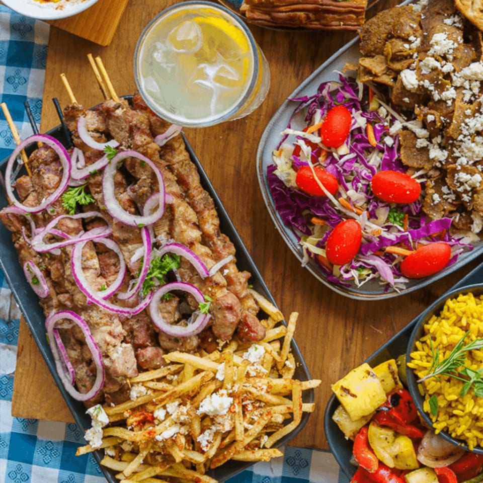 A Greek feast with skewers, fries, salad, gyro meat, rice, and grilled vegetables on a wooden table.